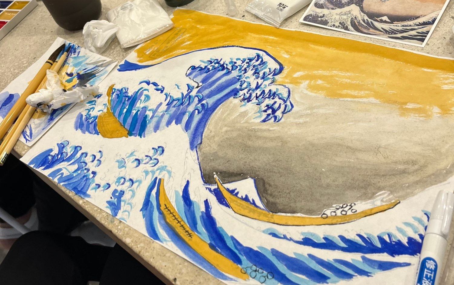 A participant’s painting of Hokusai’s The Great Wave in blue and gold pigments, with brushes and a print of the original on the table