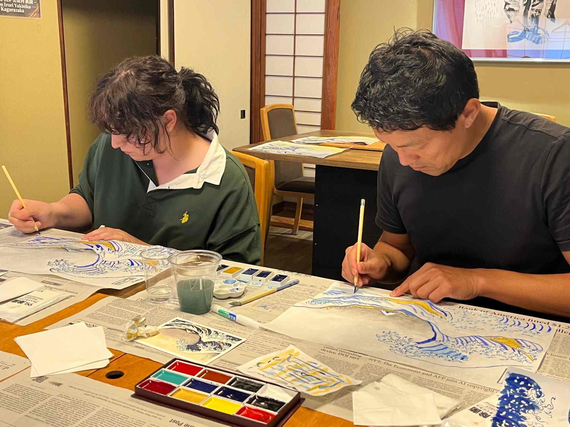 Two guests painting Hokusai’s The Great Wave with brushes and Japanese pigments at a long table